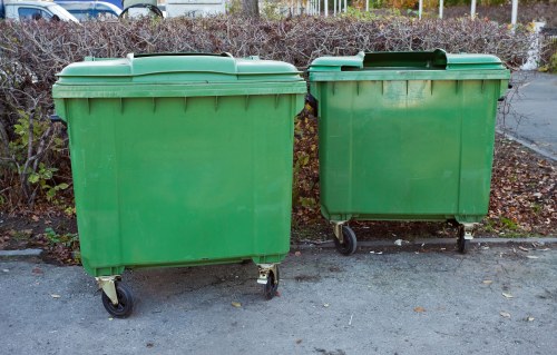 Two large green plastic wheelie bins with hinged lids are positioned side by side on a paved outdoor area, likely part of a waste management or rubbish collection service. The bin on the left appears slightly larger, with a smooth, matte finish and four small black wheels visible at the base, allowing for mobility. The bin on the right is marginally smaller, matching in style with a similar matte finish and wheels. Both bins are situated in a slightly weathered environment, with a background of low shrubbery and a gravel or asphalt surface, possibly adjacent to a property or public space within Penge, south London. The lighting suggests daytime with natural sunlight illuminating the scene, emphasizing the vibrant green color of the bins. These containers are typical for rubbish collection services around London, including local businesses or residential properties serviced by companies such as Office Clearance Penge. The overall setting indicates routine waste disposal or clearance operations, with the bins appearing clean and functional, ready for collection and outdoor use.
