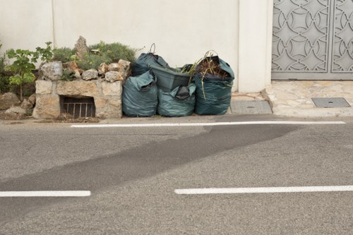 The image shows three large dark green rubbish bags filled with miscellaneous waste, placed on the pavement near the edge of a road in a residential area. The bags are made of plastic and appear to be tightly filled, with some open sections revealing tangled sticks or plant material protruding from one bag. Behind the bags is a small raised flower bed with a stone border, containing a few green plants and shrubbery. To the right, a grey decorative metal gate and part of a white wall are visible, indicating a private property. The scene is illuminated by natural daylight, casting soft shadows on the asphalt surface, which has white dashed lane markings. The overall environment suggests a typical waste collection or rubbish removal scene in Penge, close to local residential streets serviced by Office Clearance Penge's rubbish removal services.