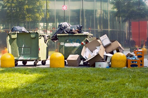 A row of three large green wheelie bins situated on a paved area next to a grassy verge, with the middle bin partially covered by a black plastic sack. To the right of the bins, there is a pile of various rubbish including opened cardboard boxes, some ripped and flattened, and discarded packaging materials. The background features a glass building reflecting nearby trees and sky, while outdoor bollards in yellow are positioned in front of the bins and rubbish pile. The scene appears to be an outdoor waste collection area, possibly associated with a local office or commercial premises, consistent with rubbish removal services in Penge. The overall setting is well-lit with natural daylight, highlighting the different textures of the trash, the smooth surface of the bins, and the vibrant green grass.