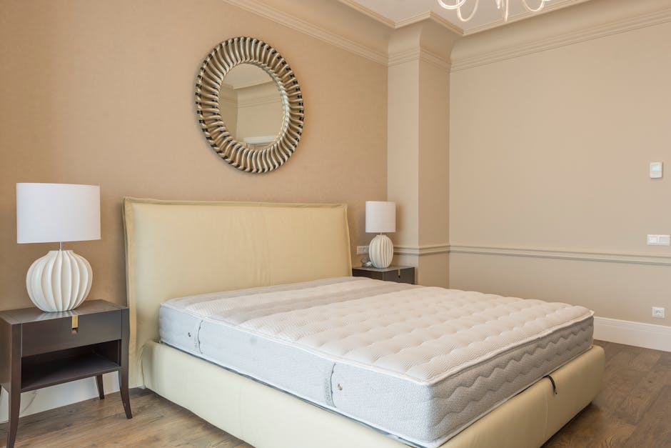 A person with long, curly dark hair, wearing a brown T-shirt and blue jeans, is seen crouching on a wooden floor while adjusting or folding a white mattress. The mattress is positioned against a white
