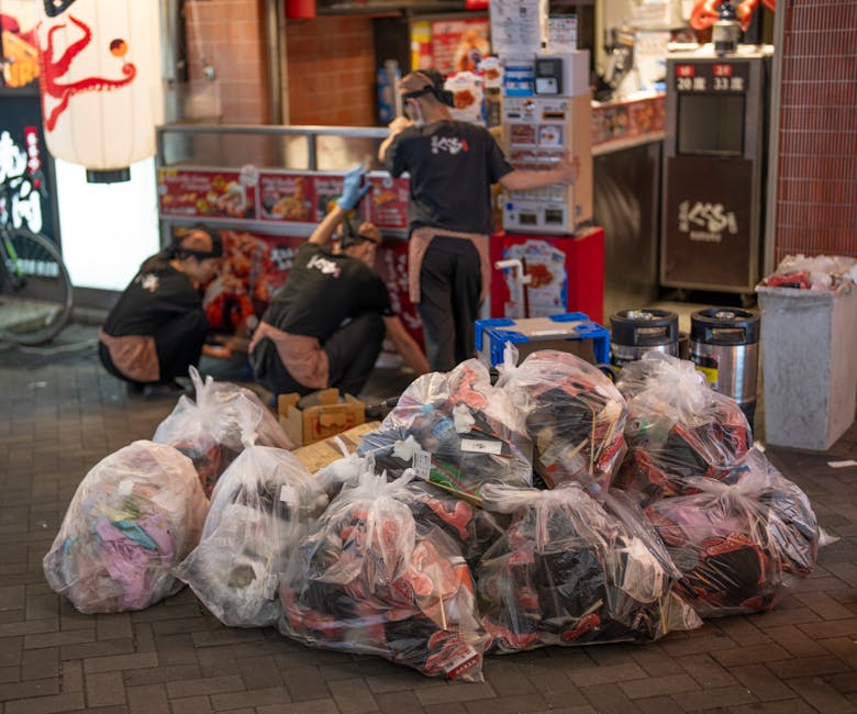 A collection of large, transparent plastic bags filled with various types of waste, including clothing and other household items, is piled on a paved sidewalk outside a commercial building at night. T